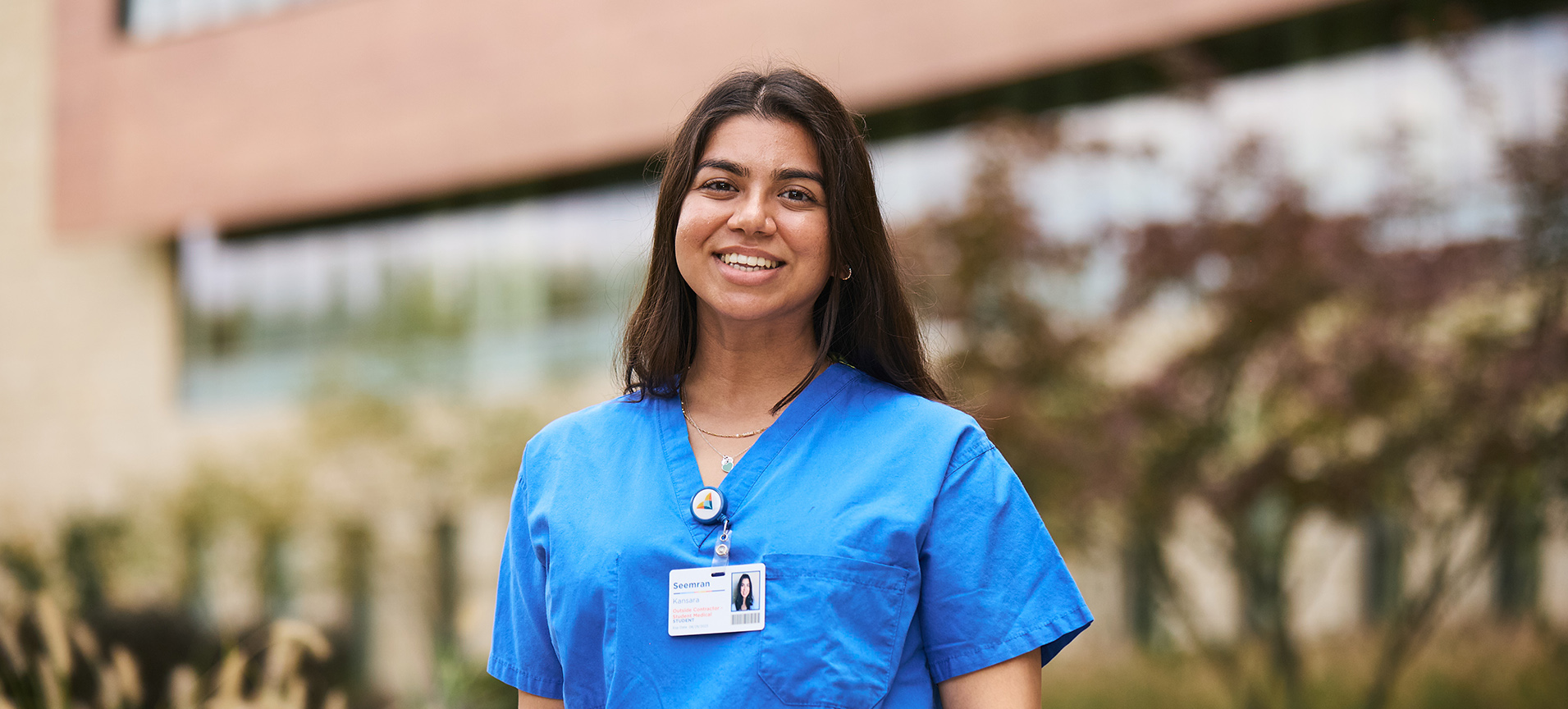 An SOM student wearing blue scrubs smiles at the camera while standing outside the Virtua Voorhees Hospital