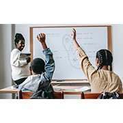 Teacher standing next to a whiteboard with two students seated at desks with their hands raised 
